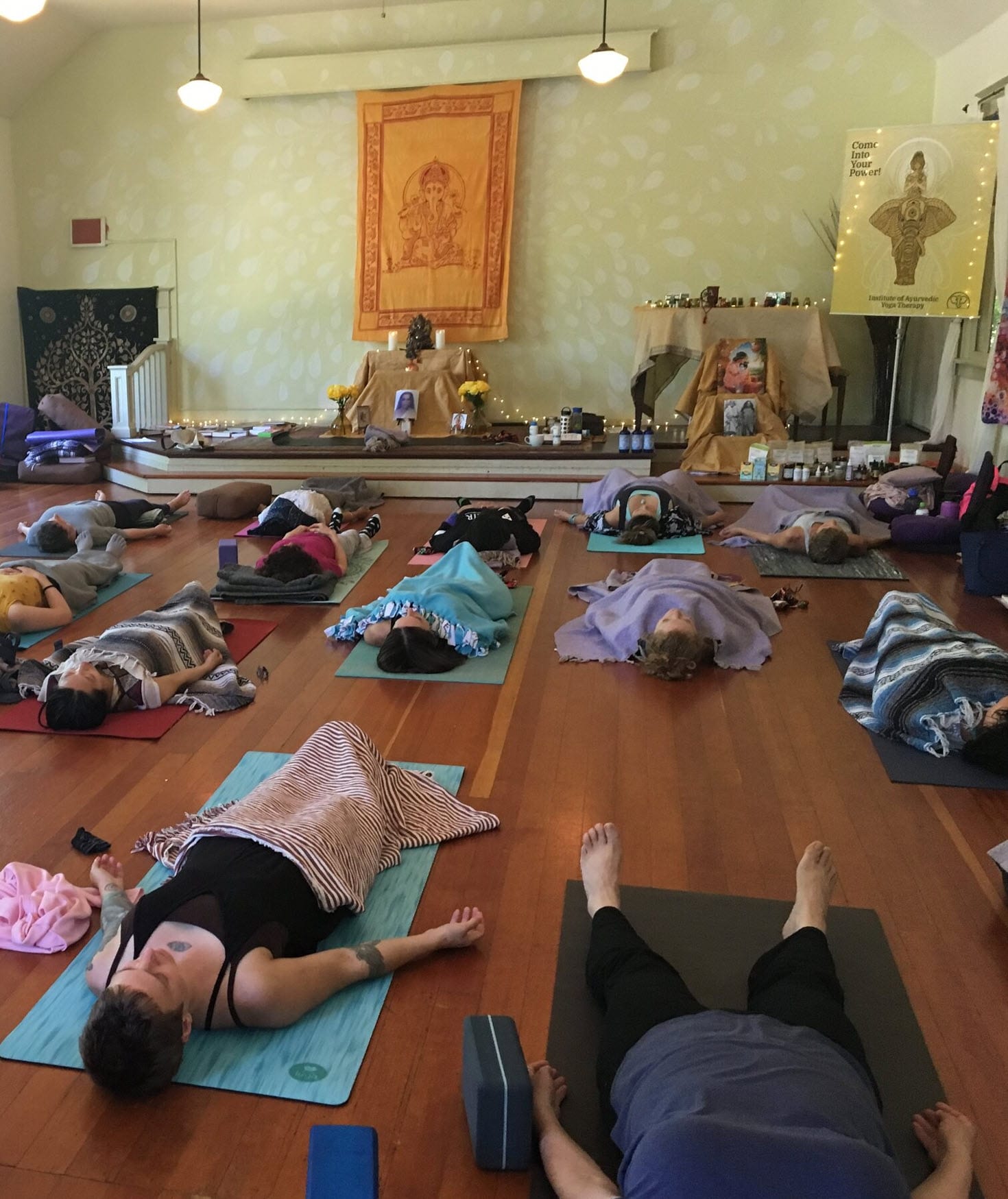 Sarasvati Foundations cohort resting in savasana on warm wood floors in the school's converted-church shala